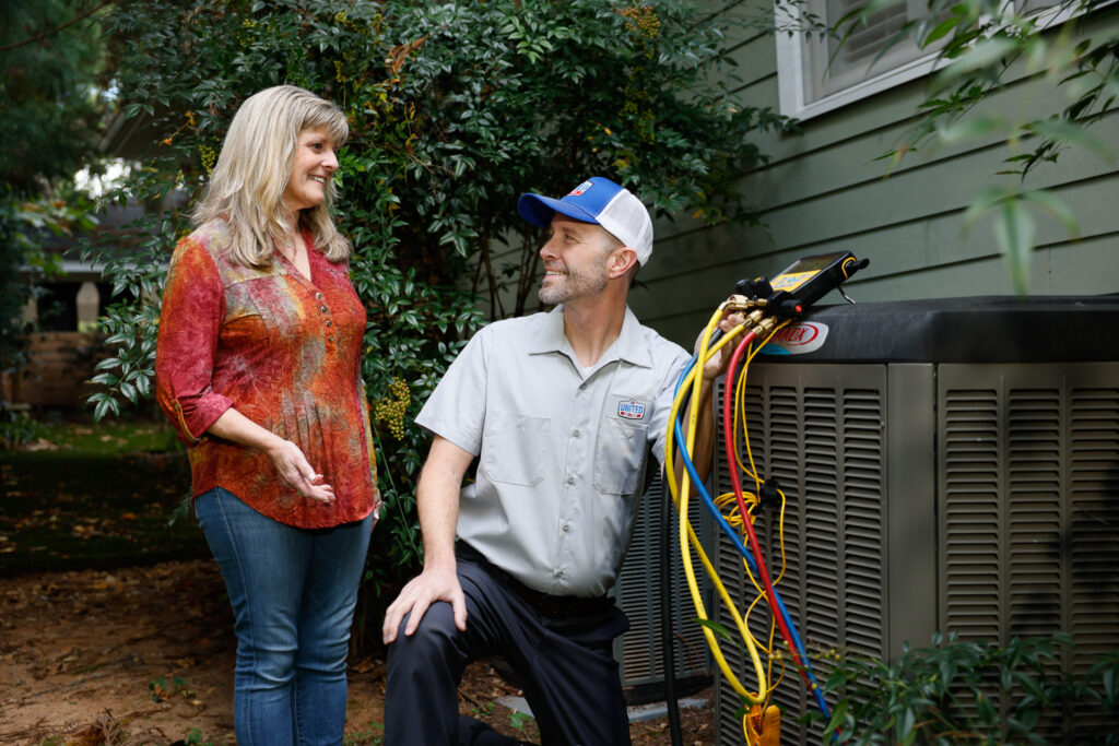 Technician speaking with homeowner while testing the HVAC system outside her green home, with tall bushes in background.