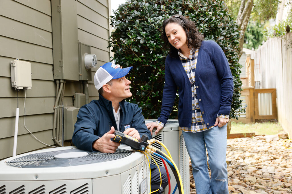Technician speaking with homeowner while working on an outdoor HVAC unit next to a gray home.