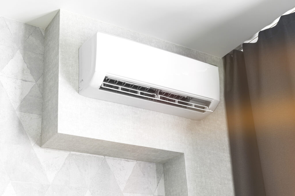 Ductless mini-split air handler mounted on a white wall in a home, next to a window with a brown curtain drawn over it.